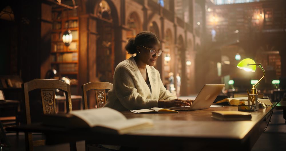 Young Woman Focused on Writing Essays and Studying for Exams Amidst Rows of Classic Books.