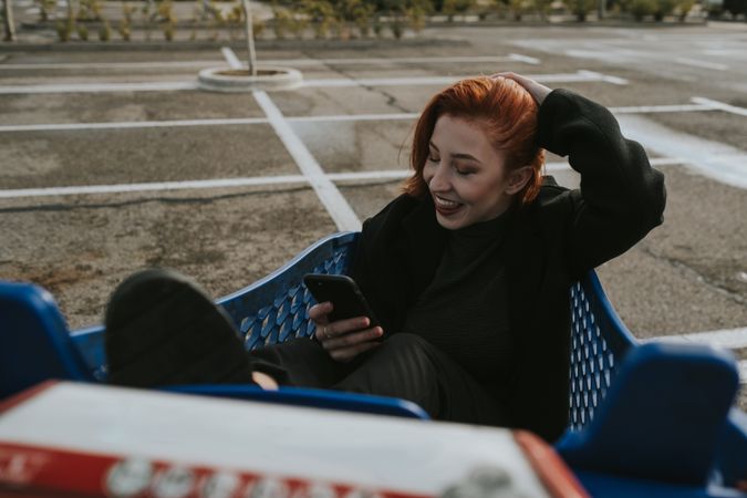 Smiling woman with smartphone in shopping trolley in parking lot