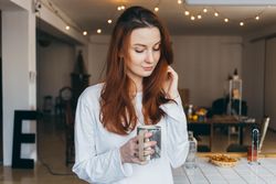 Woman plays with hair while holding a mug of a warm drink in her apartment