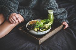 Woman in cozy sweater sitting with wooden tray of vegetarian bowl with smoothie on side