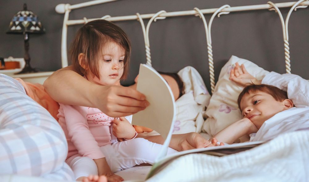 Closeup of baby girl reading book with her mother and brother in bed