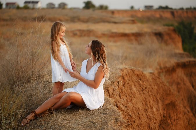 Adult female sitting looking up at standing child in summer dress on top of cliff at dusk