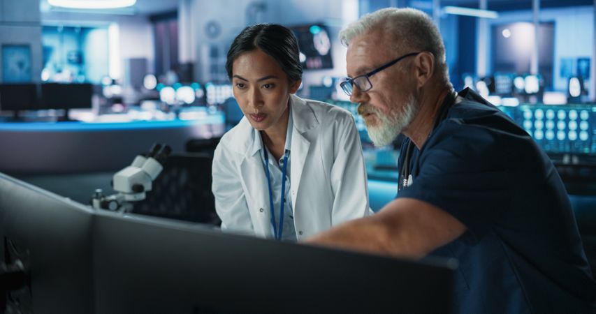 Asian Female Scientist Using Desktop Computer And Talking To Male Doctor In Modern Hospital Research Lab.