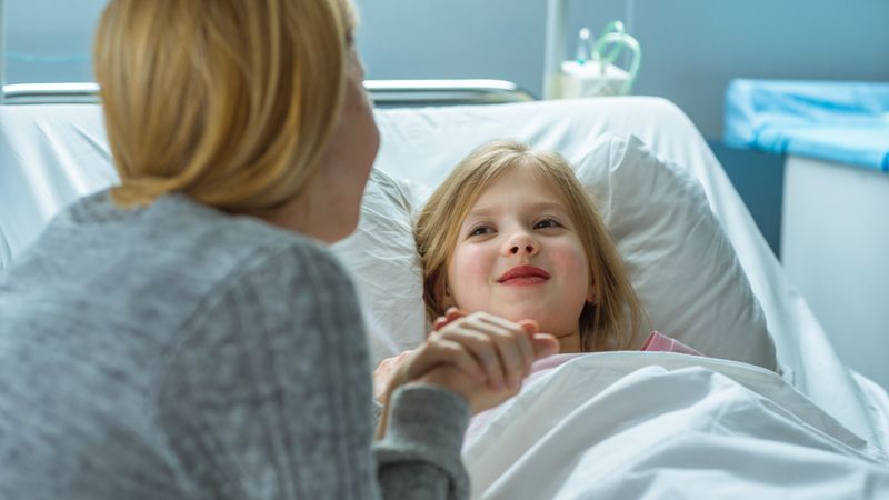 Cute Little Girl Lies on a Bed in the Children's Hospital and Talking with Her Mother Sits Beside