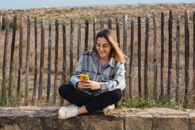Young female millennial resting on stone border while using mobile phone at roadside