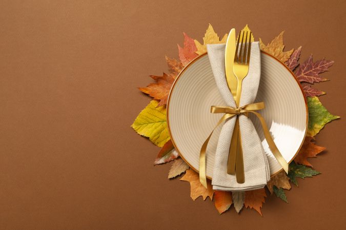 Golden cutlery on plate and leaves on brown background, space for text