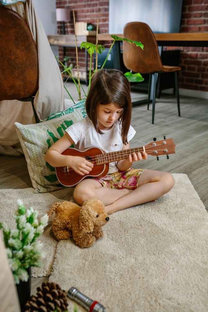 Little girl playing ukulele sitting on carpet next to handmade shelter tent in living room at home
