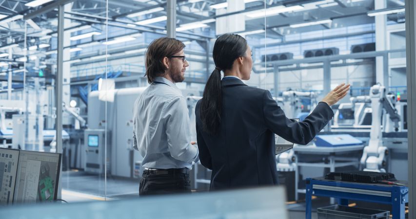 Young Engineer and Asian Technician Having Conversation and Using a Laptop Computer. Specialists Monitoring, Analyzing Conditions at a Modern Electronics Factory with Automated Conveyor