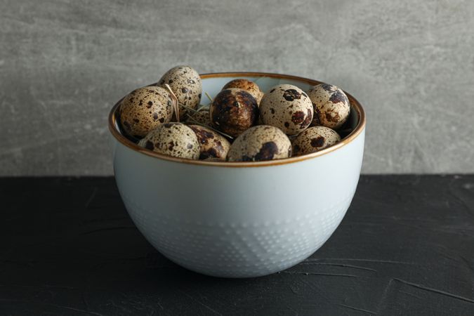 Quail eggs in bowl on dark table against grey background, space for text
