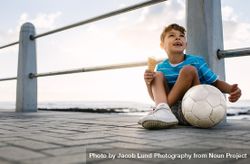 Happy boy eating an ice cream sitting on a pier with soccer ball 4BaBBx