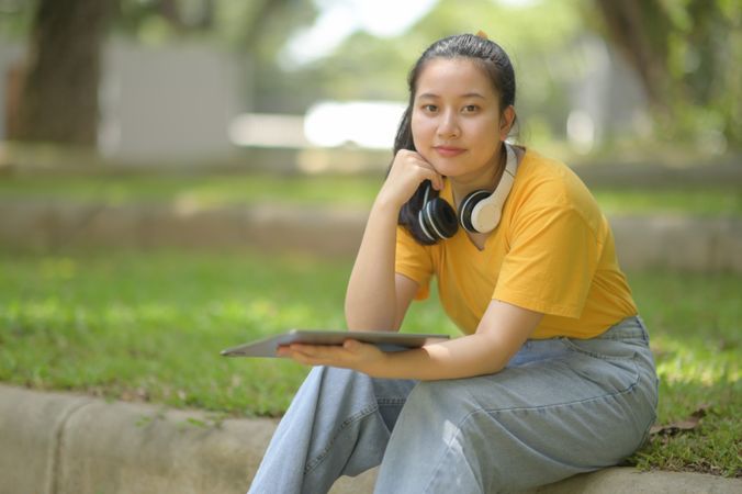 Woman in the yellow shirt wearing headphones on the neck, holding tablet sitting in the garden looking at the camera.