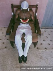 Topless man in light pants holding a slice of watermelon sitting wooden armchair 5lMom4