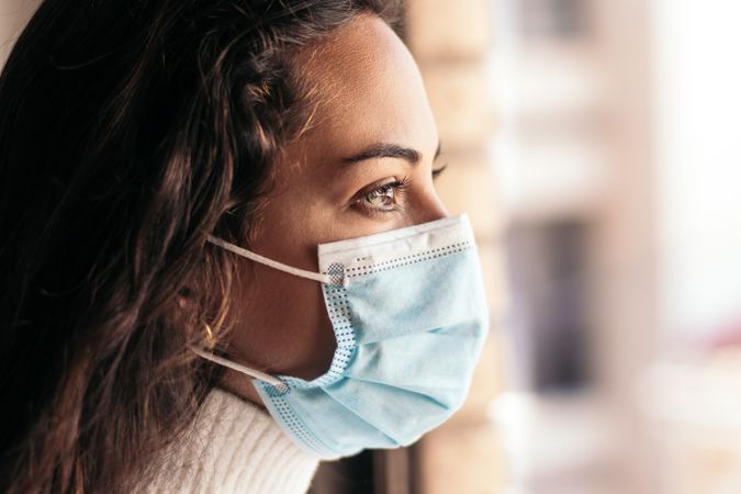 Young woman wearing face mask looking out the window at home