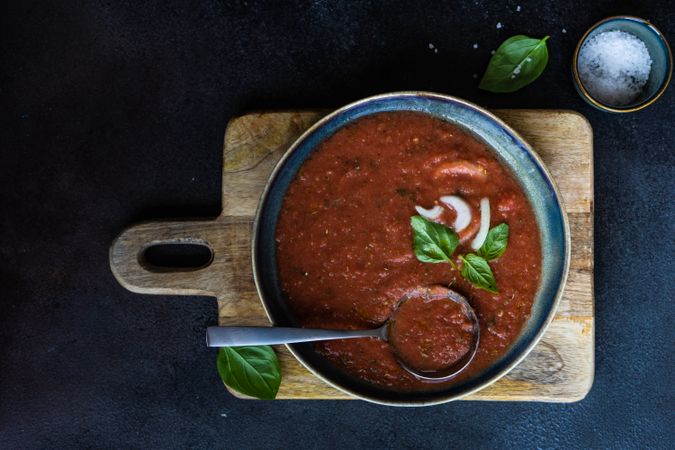 Top view of large bowl & spoon in Spanish gazpacho with basil leaf garnish and salt in blue bowl