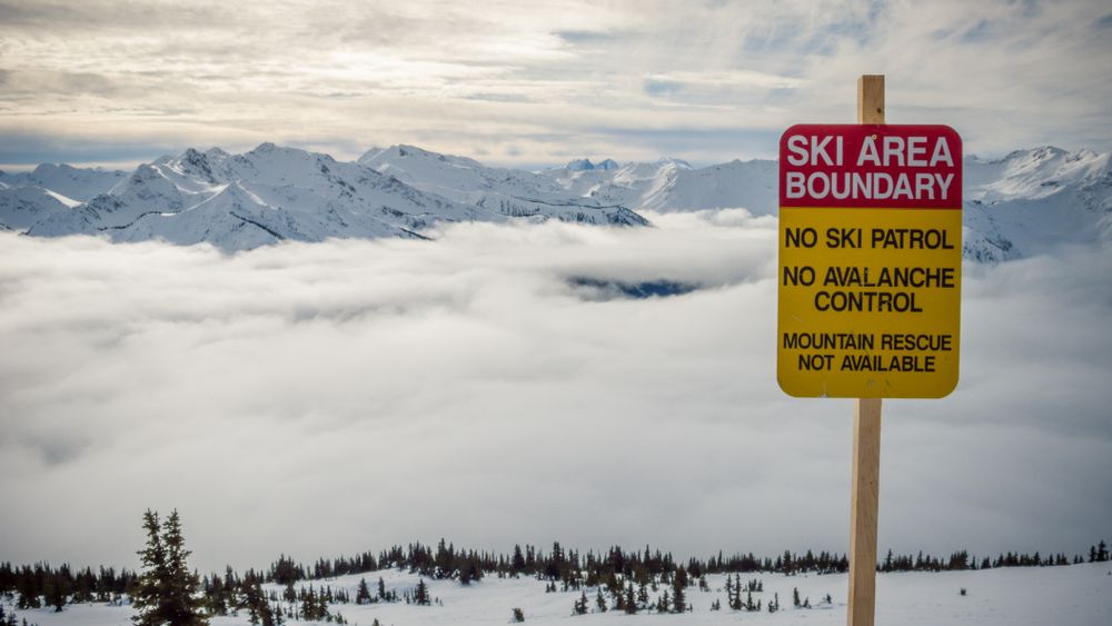 Sign with ski area boundary title on hill with snow and clouds between mountains