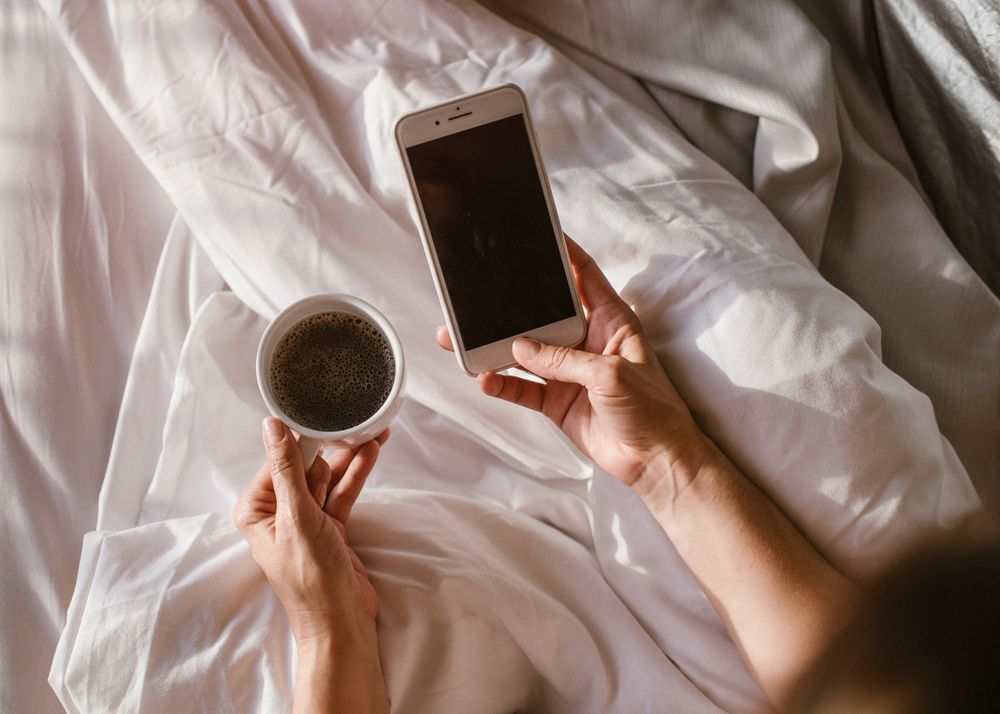 Woman having hot drink and using smartphone in bed