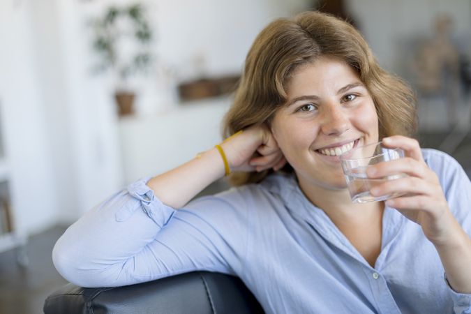 Front view portrait of a happy woman holding a glass of water sitting on a couch at home