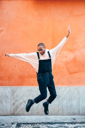 Male in overalls and hoodie jumping while looking down in front of wall
