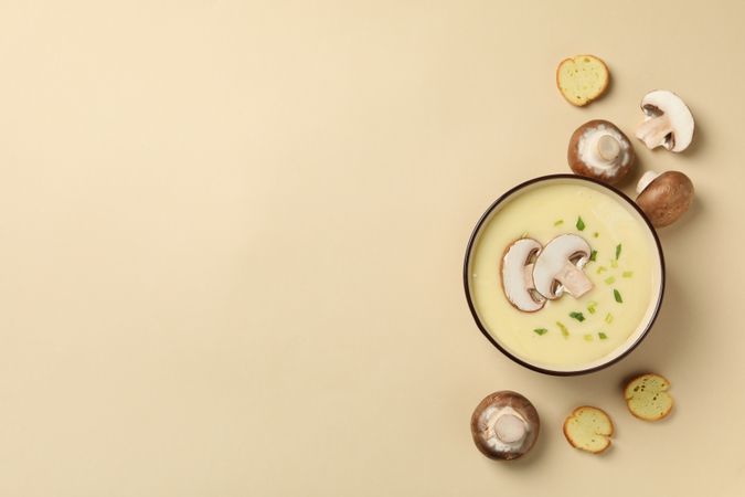 Top view of mushrooms surrounding bowl of soup on beige background