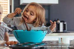 Little girl licking spoon while mixing batter for baking in kitchen  and her brother standing by 0LLXV0
