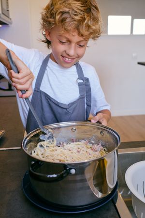 Crop smiling boy stirring spaghetti with carbonara sauce in kitc