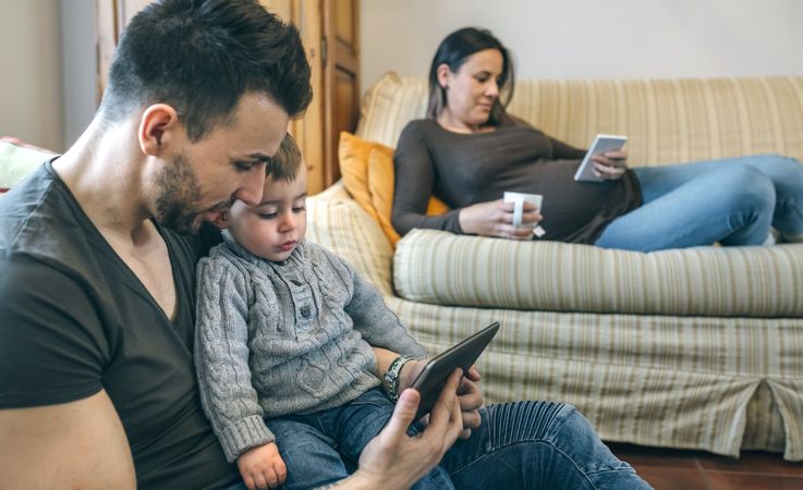 Father looking tablet with little son while pregnant mother looks mobile