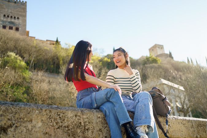 Two female friends sitting on stony wall on nice day in Spain