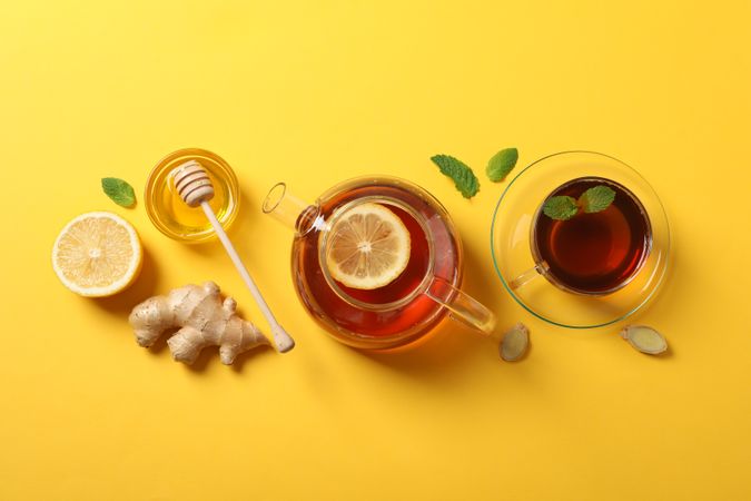Flat lay. Cup of tea, teapot, lemon, ginger, mint, honey and dipper on yellow background, copy space