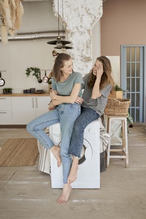 Laughing girlfriends sitting on washing machine in house