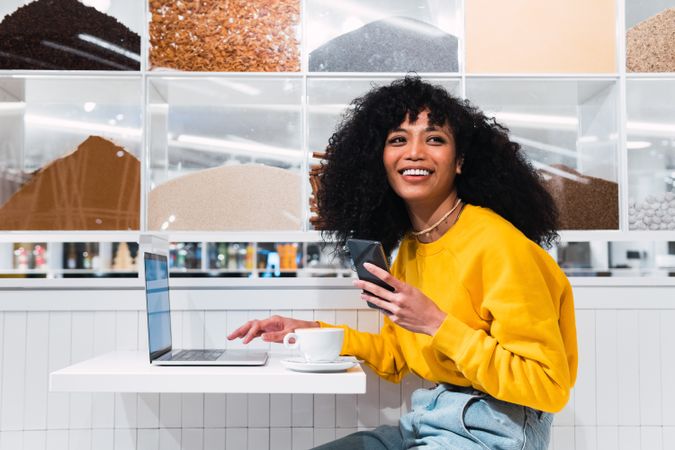Smiling woman with phone using laptop in cafe