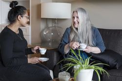 Two women sit on a couch smiling with cups of tea