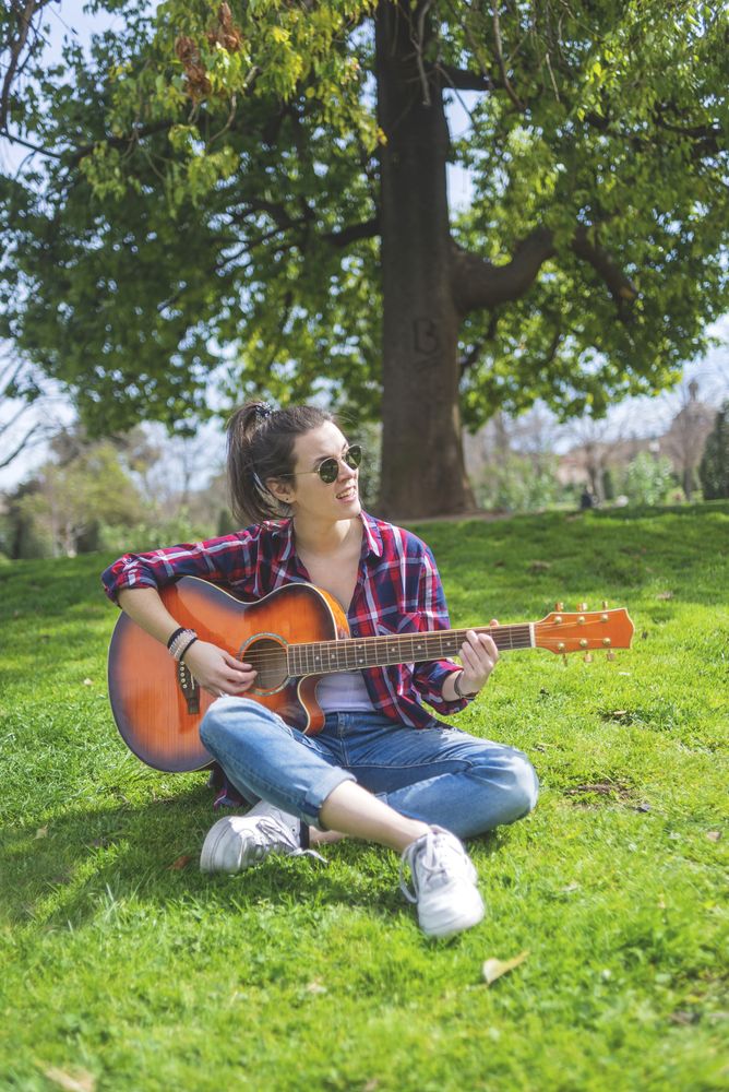 Front view of a young hipster woman wearing sunglasses, sitting on grass in a park while enjoying playing guitar