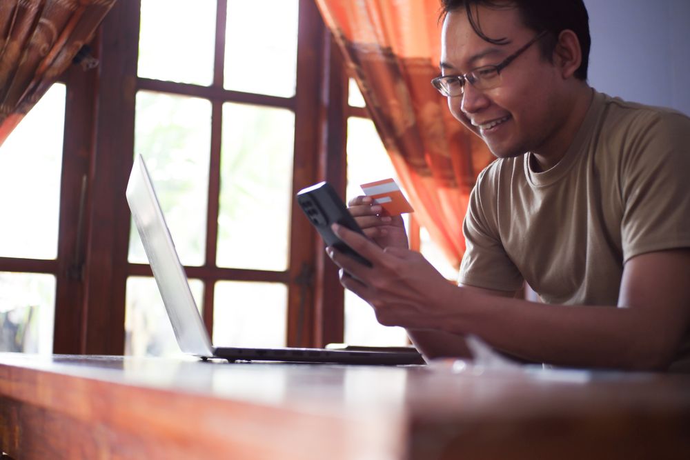 Asian Male with shirt doing online transaction in living room with credit card smartphone and laptop