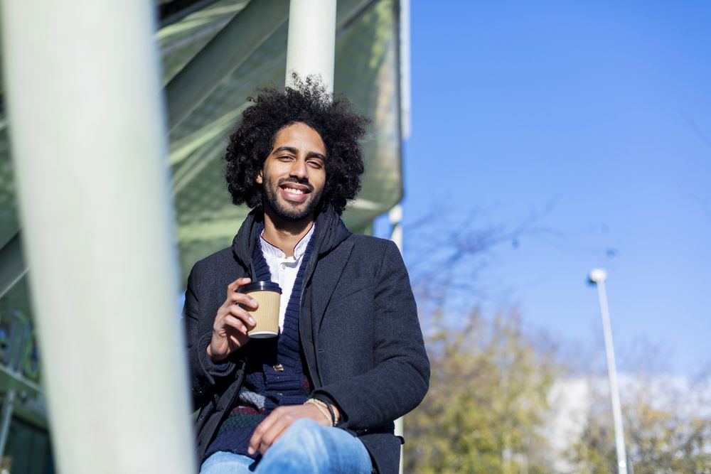 Happy Black man in jacket with warm beverage balancing between poles outside