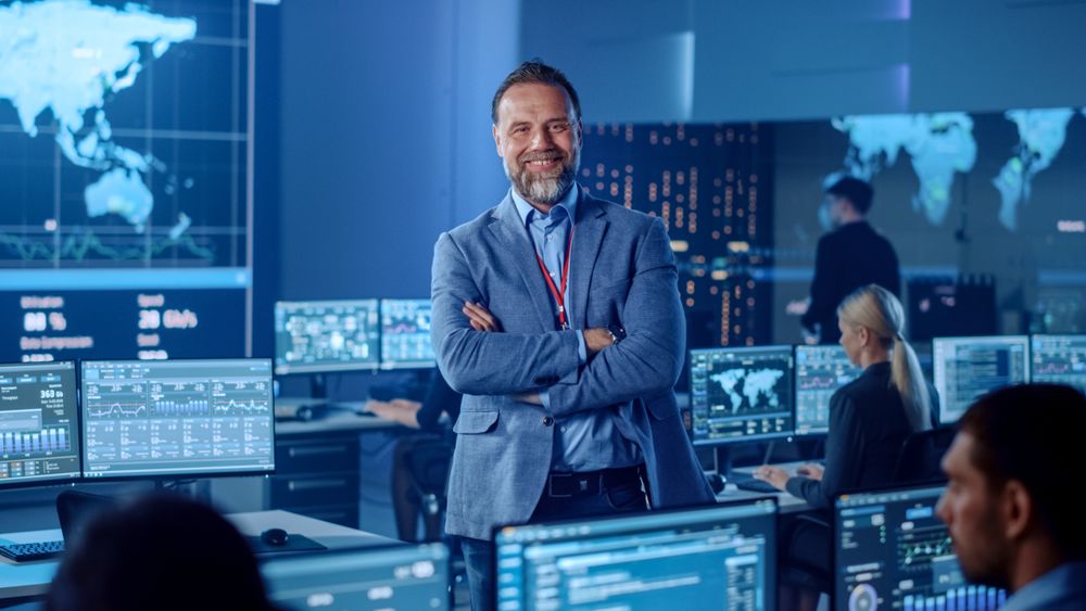 Confident Executive Stands in High-tech Control Room. Team Monitors Global Data on Screens. Blue-lit Environment With World Map Displays. Advanced Technology for Real-time Analysis.