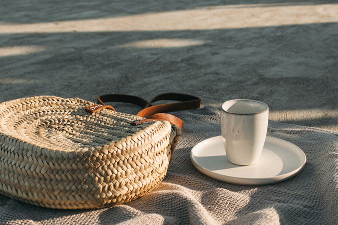 Elegant wicker straw bag on beige towel in sunlight with cup of coffee on plate