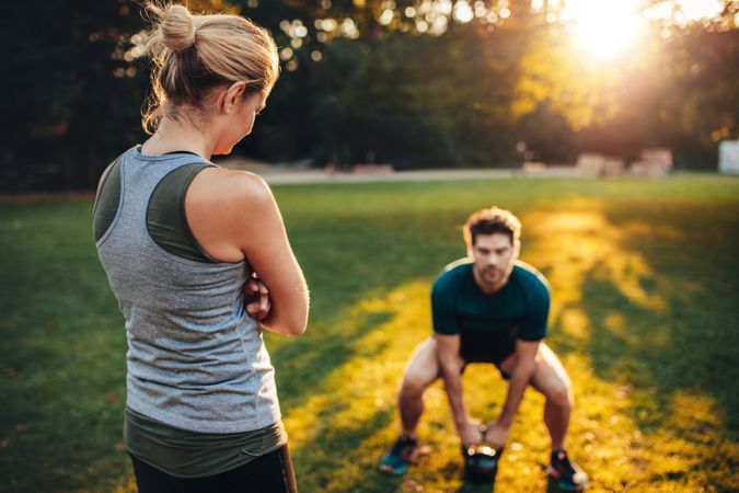 Female trainer standing in the park with man doing weight training with kettlebell