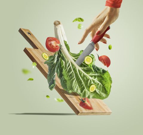 Women hand with knife , flying salad ingredients and cutting board at light green background. Creative food levitation. Healthy eating and dieting