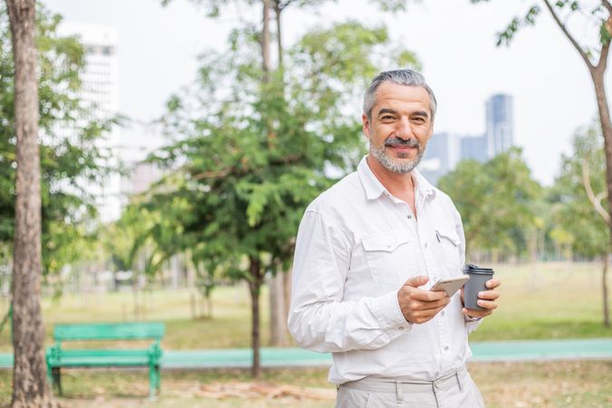 Content grey haired man standing in a park with phone and to-go coffee