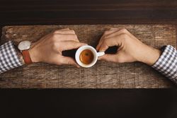 Man wearing plaid shirt and watch holding espresso shot on wooden board