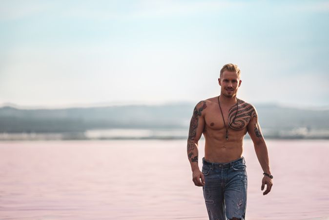 Undressed muscular man walking on shore of pink lake
