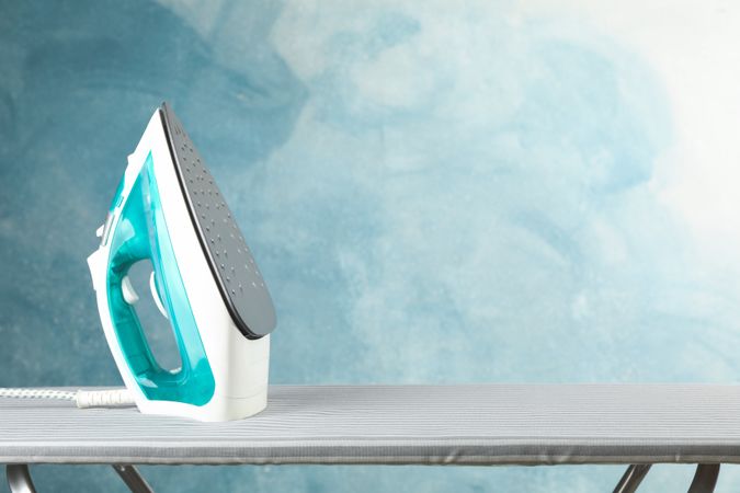 Iron on ironing board against blue background, space for text