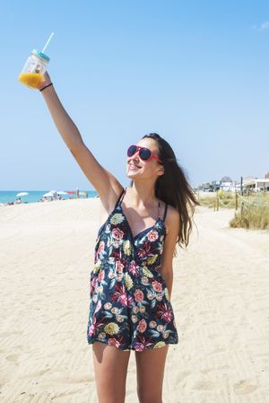A dark haired happy woman in dress and sunglasses, holding out a delicious juice drink in her hand