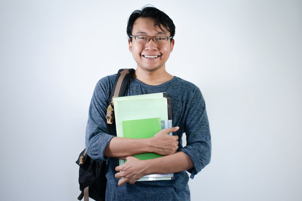 Asian young male student wearing bag and carrying books smiling and happy expression
