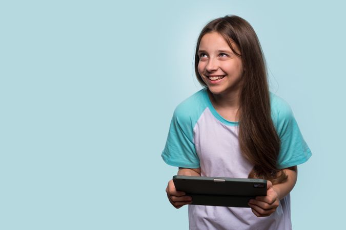 Cheerful girl looking upward, holding tablet in relaxed stance.