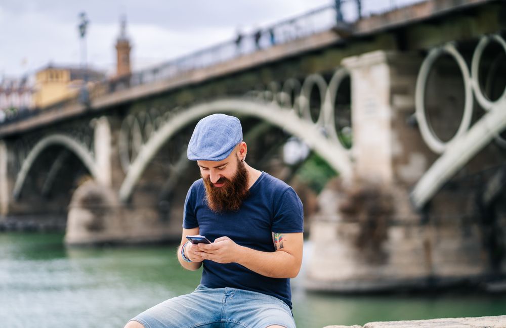 Bearded young man in casual wear using smartphone while walking on quay