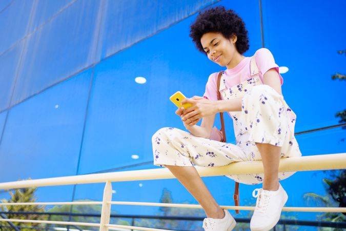 Woman sitting on handrails in front of reflective building texting on phone