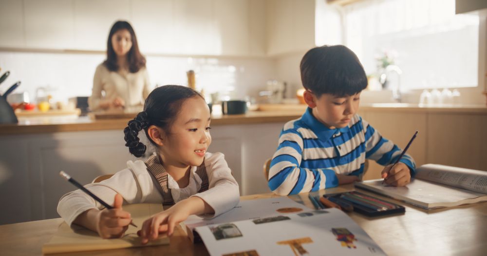 Portrait of a Little Korean Kids Sitting at a Kitchen Table at Home in the Morning Mother is Cooking in the Background.