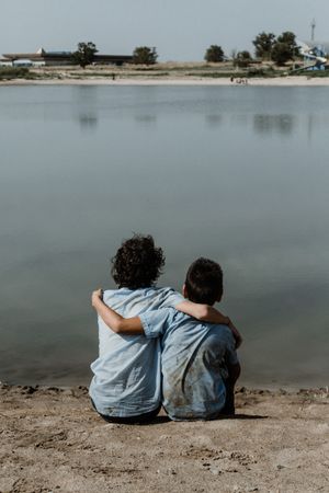 Unrecognizable kids sitting on ground by pond