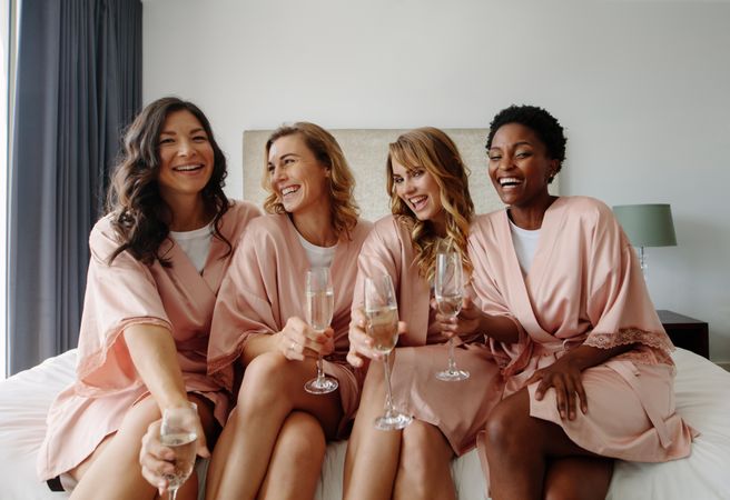 Females in sleeping robe sitting on bed and having champagne